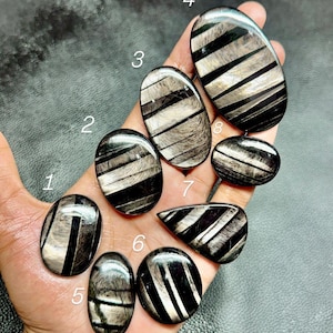 Puede incluir: Una colección de ocho piedras preciosas pulidas, de forma ovalada y de lágrima, con rayas negras y plateadas, etiquetadas con los números del uno al ocho. Cada piedra presenta un patrón de bandas negras alternas e inclusiones fibrosas plateadas.