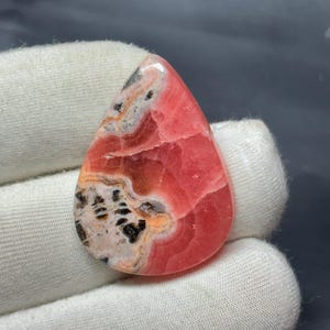 以下が含まれることがあります： 鮮やかなピンク色の涙型の宝石。白、黒、オレンジの複雑な模様が特徴です。研磨された表面は光を反射し、石の自然なバリエーションを際立たせています。ジュエリーや装飾に適しています。