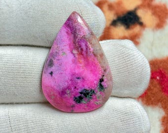 May include: A teardrop-shaped gemstone with a vibrant pink hue, accented by darker pink, purple, and green inclusions. The polished stone is set against a white fabric background, showcasing its natural beauty and unique color variations.