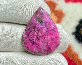 May include: A teardrop-shaped gemstone with a vibrant pink hue, veined with darker purple and flecks of green. The polished stone is held in a white glove, showcasing its smooth surface and natural patterns. The gemstone is a unique piece of jewelry.