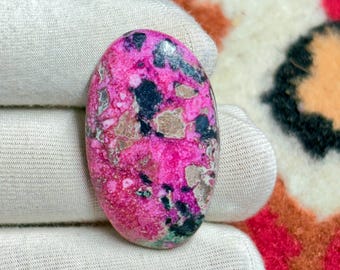 May include: An oval-shaped gemstone with a vibrant pink base color, speckled with black, green, and tan inclusions. The polished surface of the stone reflects light, showcasing its unique pattern. The gemstone is held in a white glove, highlighting its natural beauty.