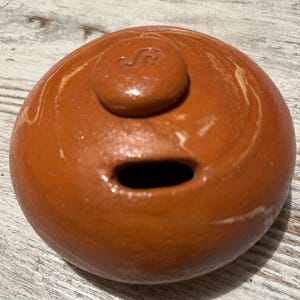 Può includere: Un oggetto rotondo in ceramica color terracotta con un piccolo elemento circolare rialzato sulla parte superiore. L'elemento superiore presenta un piccolo disegno stilizzato. Un'apertura scura di forma ovale è visibile sul lato. La superficie ha un effetto marmorizzato.