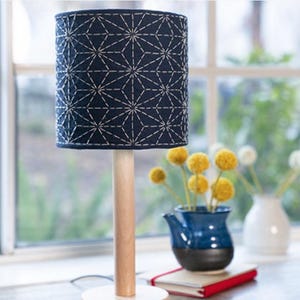 May include: A navy blue table lamp with a geometric white stitched pattern on the lampshade. The lamp has a light wood base and a white circular base. The lamp is on a table with a vase of yellow flowers.