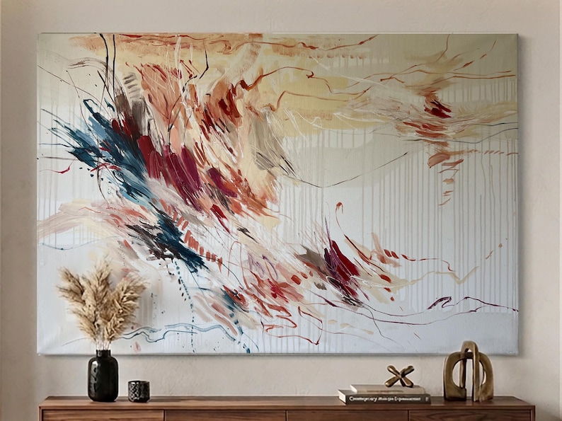 May include: Abstract canvas art with a cream background and vertical white lines. The artwork features dynamic brushstrokes in shades of red, blue, and beige. A black vase with dried flowers and decorative items are on a wooden surface.