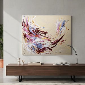May include: Abstract canvas art with a beige background and dynamic brushstrokes in shades of burgundy, pink, blue, and white. The artwork is mounted on a wall above a dark wood cabinet.