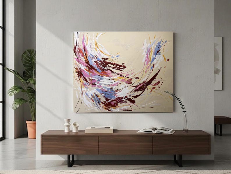 May include: Abstract canvas art with a beige background and dynamic brushstrokes in shades of burgundy, pink, blue, and white. The artwork is mounted on a wall above a dark wood cabinet.
