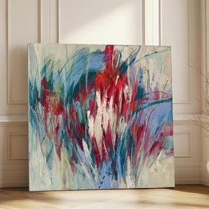 May include: Large square abstract painting with a vibrant display of colors. The artwork features bold brushstrokes in shades of red, blue, and teal, creating a dynamic and energetic composition. The painting is displayed on a wooden floor.