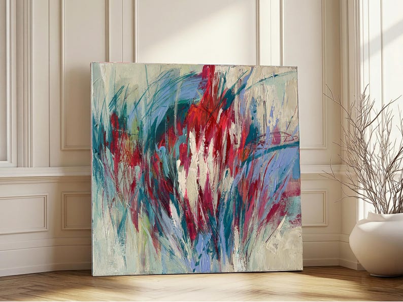 May include: Large square abstract painting with a vibrant display of colors. The artwork features bold brushstrokes in shades of red, blue, and teal, creating a dynamic and energetic composition. The painting is displayed on a wooden floor.