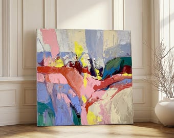 Puede incluir: Una gran pintura abstracta cuadrada con una paleta de colores vibrantes. La obra de arte presenta amplias pinceladas en tonos de rosa, amarillo, azul, rojo y verde. La pintura está apoyada contra una pared blanca con suelo de madera.