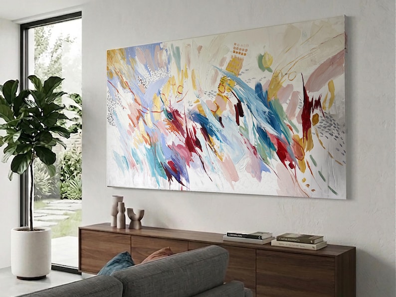 Puede incluir: Gran pintura abstracta con fondo blanco y pinceladas coloridas en tonos azules, rojos, amarillos y rosas. La obra de arte est&aacute; montada en una pared blanca sobre un mueble de madera.