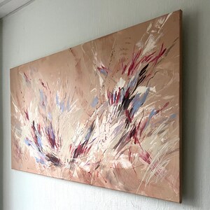 May include: Abstract painting on canvas with a beige background and dynamic brushstrokes in white, red, and blue. The artwork is mounted on a wooden frame and leans against a light gray wall.