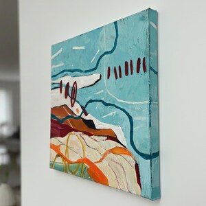 May include: Square abstract painting with a textured surface. The artwork features a light blue background with white, orange, and dark red shapes. The painting is mounted on a canvas frame and displayed on a white wall.