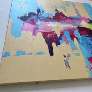 May include: Abstract painting on canvas with a textured, beige background. Bold brushstrokes in shades of blue, pink, purple, red, and green create a dynamic composition. The artwork is mounted on a wooden frame.