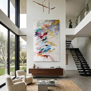 Puede incluir: Sal&oacute;n moderno con grandes ventanales, dos sillones color crema y una alfombra rectangular tejida. Una gran pintura abstracta con pinceladas rojas, azules y amarillas cuelga sobre un mueble de madera oscura. Una mesa de centro de cristal en el centro.