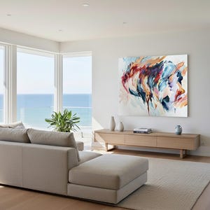 May include: A modern living room with a beige sectional sofa, a light wood cabinet, and a large abstract painting. The painting features a colorful portrait. The room has large windows with a view of the ocean.