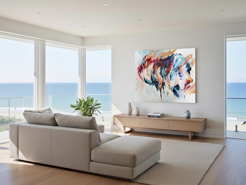 May include: A modern living room with a beige sectional sofa, a light wood cabinet, and a large abstract painting. The painting features a colorful portrait. The room has large windows with a view of the ocean.