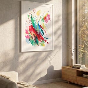 May include: A framed vertical wall art print featuring an abstract watercolor composition with vibrant brushstrokes in red, green, blue, and yellow on a white background, displayed in a simple white frame mounted on a textured concrete wall.