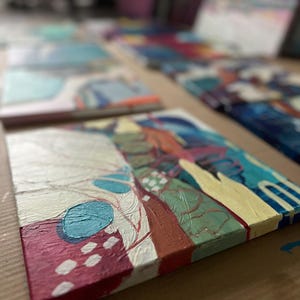 May include: Abstract acrylic paintings on canvas. The artwork features a mix of colors, including red, white, blue, and yellow. The paintings are displayed on a brown surface, with several other paintings in the background.