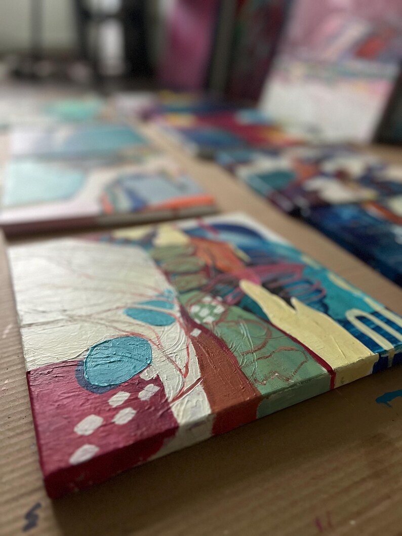 May include: Abstract acrylic paintings on canvas. The artwork features a mix of colors, including red, white, blue, and yellow. The paintings are displayed on a brown surface, with several other paintings in the background.