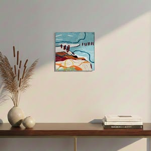 May include: Abstract landscape painting with a blue sky, brown and red hills, and orange and yellow accents. The artwork is displayed on a white wall above a wooden table with a vase of dried plants and books.