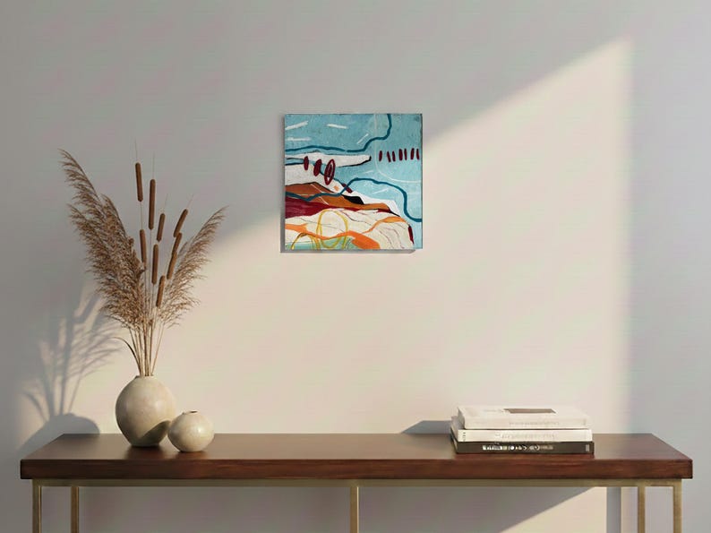 May include: Abstract landscape painting with a blue sky, brown and red hills, and orange and yellow accents. The artwork is displayed on a white wall above a wooden table with a vase of dried plants and books.