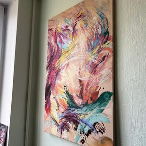 以下が含まれることがあります： ベージュの背景に抽象的な絵画。ピンク、紫、黄色、ティール、白の色合いでダイナミックな筆致が特徴です。絵画は木製のフレームに取り付けられ、淡い緑色の壁に掛けられています。