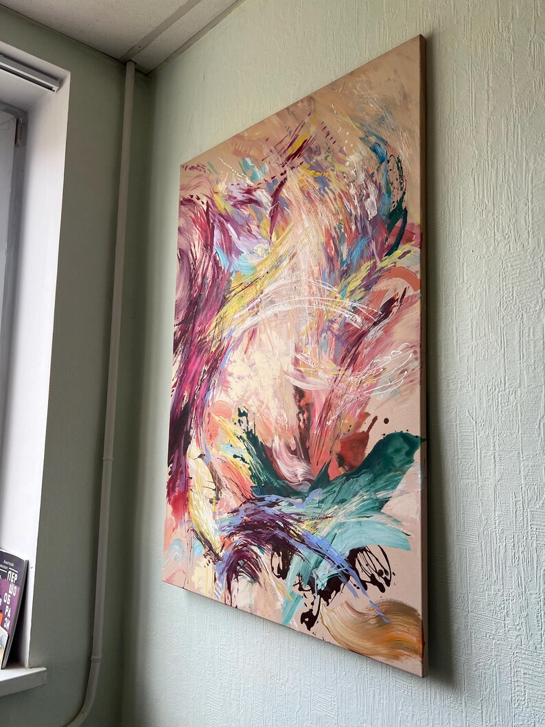 以下が含まれることがあります： ベージュの背景に抽象的な絵画。ピンク、紫、黄色、ティール、白の色合いでダイナミックな筆致が特徴です。絵画は木製のフレームに取り付けられ、淡い緑色の壁に掛けられています。