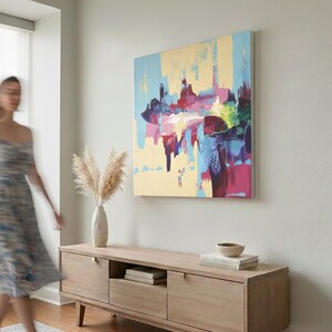 May include: Abstract art painting with a light yellow background and colorful brushstrokes of blue, pink, and purple. The artwork hangs above a wooden cabinet with a vase of pampas grass.