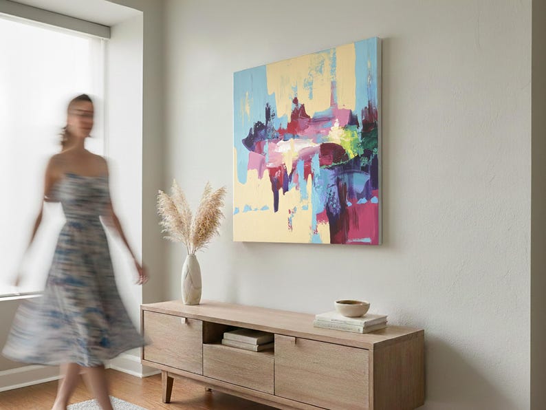 May include: Abstract art painting with a light yellow background and colorful brushstrokes of blue, pink, and purple. The artwork hangs above a wooden cabinet with a vase of pampas grass.