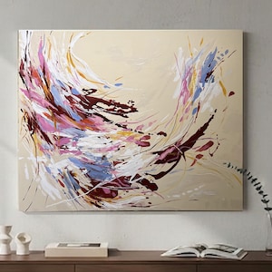 May include: Abstract painting on canvas with a beige background. The artwork features dynamic brushstrokes in shades of pink, white, blue, burgundy, and yellow. The painting is mounted on a wall above a wooden surface.