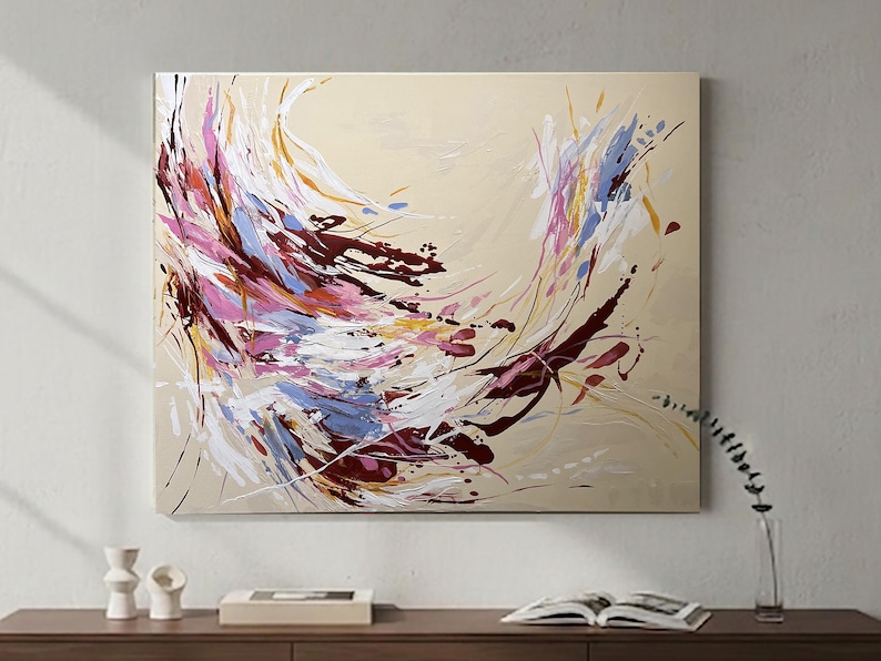 May include: Abstract painting on canvas with a beige background. The artwork features dynamic brushstrokes in shades of pink, white, blue, burgundy, and yellow. The painting is mounted on a wall above a wooden surface.