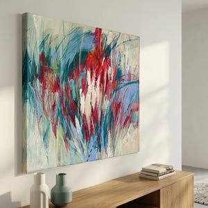 May include: Large square abstract painting with a cream background and bold strokes of red, blue, and green. The artwork is mounted on a white wall above a wooden cabinet. Two vases and a stack of books are on the cabinet.
