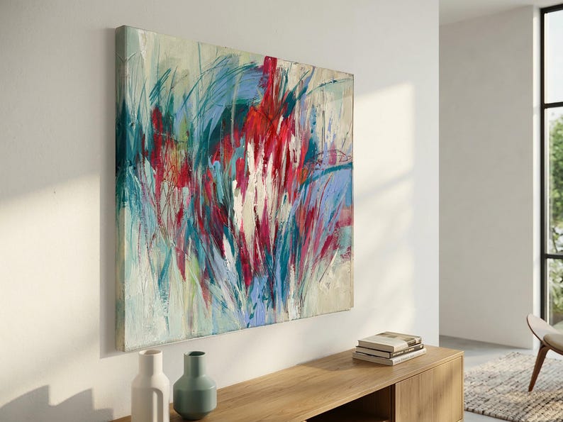 May include: Large square abstract painting with a cream background and bold strokes of red, blue, and green. The artwork is mounted on a white wall above a wooden cabinet. Two vases and a stack of books are on the cabinet.