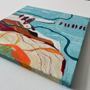 May include: Abstract painting on a square canvas. The artwork features a textured surface with a palette of colors including light blue, orange, red, and white. The composition includes lines and shapes, creating a modern art piece.