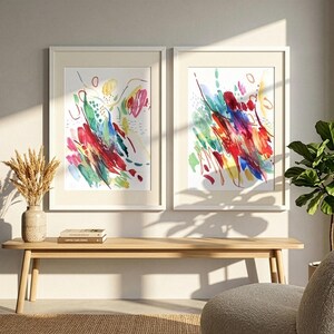 May include: Two framed vertical wall art prints featuring abstract watercolor brushstrokes in vibrant shades of red, blue, yellow, and green on a white background, displayed side by side in light-colored wooden frames on a plain wall.