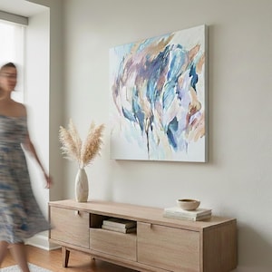 May include: An abstract painting with blue, purple, and tan brushstrokes hangs above a light wood cabinet. A vase of dried pampas grass and a small bowl sit on the cabinet. A person in a dress is blurred in the foreground.