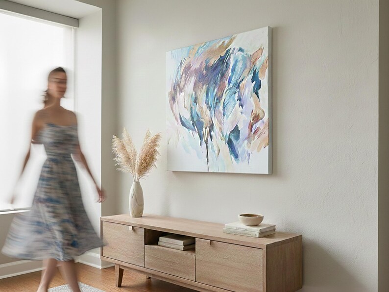 May include: An abstract painting with blue, purple, and tan brushstrokes hangs above a light wood cabinet. A vase of dried pampas grass and a small bowl sit on the cabinet. A person in a dress is blurred in the foreground.