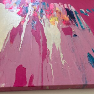 May include: Abstract painting on canvas with a vibrant pink background. Silver, white, and various shades of pink and blue paint drip down from the top, creating a dynamic and textured effect. The artwork has a modern and expressive style.