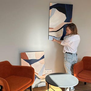 May include: An abstract painting with a vertical orientation, featuring shapes in shades of white, navy, and peach. The artwork is being hung on a gray wall. Two orange armchairs and a small round table are in the foreground.