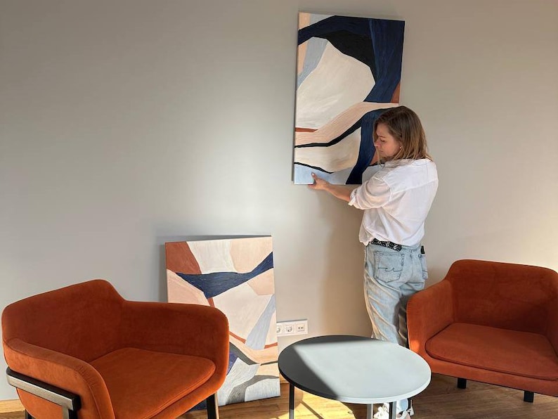 May include: An abstract painting with a vertical orientation, featuring shapes in shades of white, navy, and peach. The artwork is being hung on a gray wall. Two orange armchairs and a small round table are in the foreground.