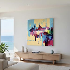 May include: Abstract art painting with a gold background and splashes of blue, pink, and purple. The artwork is displayed on a white wall above a wooden cabinet. The room has large windows overlooking the ocean.