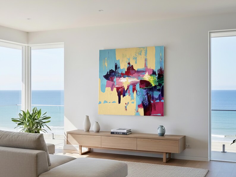 May include: Abstract art painting with a gold background and splashes of blue, pink, and purple. The artwork is displayed on a white wall above a wooden cabinet. The room has large windows overlooking the ocean.