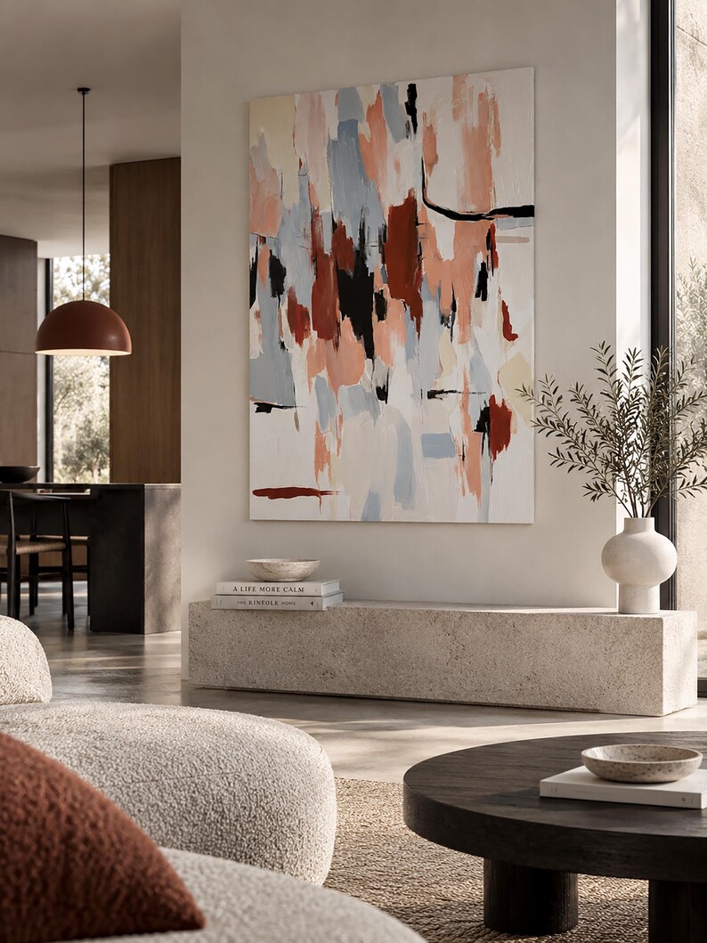 May include: A large rectangular canvas painting featuring abstract brushstrokes in shades of peach, light blue, cream, deep red, and black. The artwork is displayed on a wall above a long, low, textured stone bench in a neutral-toned room.