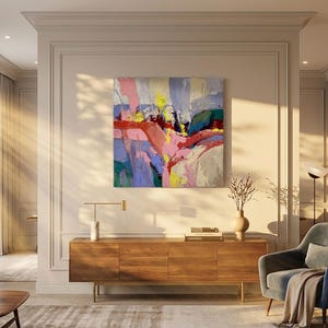 May include: An abstract artwork with pink, yellow, blue, and red strokes hangs above a wooden cabinet. A blue armchair with a blanket is to the right. A vase with dried branches and a modern lamp are on the cabinet.