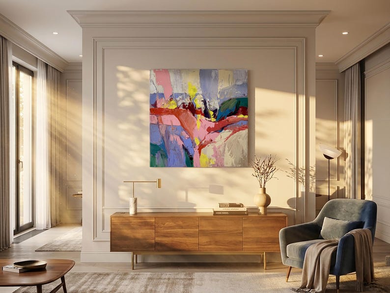 May include: An abstract artwork with pink, yellow, blue, and red strokes hangs above a wooden cabinet. A blue armchair with a blanket is to the right. A vase with dried branches and a modern lamp are on the cabinet.