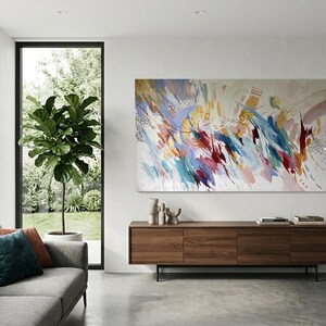 Puede incluir: Pintura de arte abstracto con una vibrante mezcla de colores, incluyendo azul, rojo, amarillo y blanco. La obra de arte se exhibe sobre un mueble de madera en un sal&oacute;n moderno. La habitaci&oacute;n tambi&eacute;n cuenta con un sof&aacute; gris, una ventana grande y una planta en maceta.