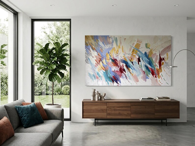 Puede incluir: Pintura de arte abstracto con una vibrante mezcla de colores, incluyendo azul, rojo, amarillo y blanco. La obra de arte se exhibe sobre un mueble de madera en un sal&oacute;n moderno. La habitaci&oacute;n tambi&eacute;n cuenta con un sof&aacute; gris, una ventana grande y una planta en maceta.