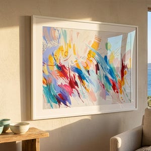 May include: A large rectangular framed wall art piece featuring an abstract composition with vibrant brushstrokes in shades of blue, red, yellow, and pink against a light cream background, displayed in a clean white frame on a textured wall.