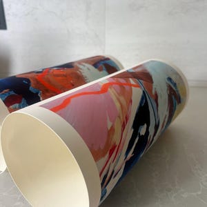 Puede incluir: Dos rollos de papel cil&iacute;ndricos con impresiones de arte abstracto. Los dise&ntilde;os muestran una paleta de colores vibrantes de naranja brillante, rosa suave, azul marino profundo y crema. Los patrones de pinceladas est&aacute;n superpuestos.