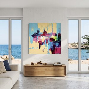 May include: An abstract painting with a light yellow background and colorful brushstrokes of blue, pink, red, and green. The artwork hangs above a wooden cabinet in a bright, airy room with a view of the ocean.
